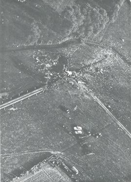Zwart-wit luchtfoto van een heuvelachtig landschap met wegen, een kruispunt en een klein gebouw.