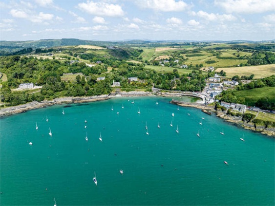Luchtfoto van een turquoise baai met zeilboten, omringd door groene heuvels en kustwoningen.