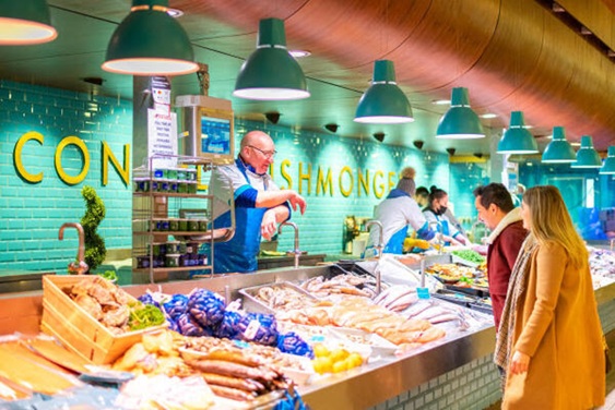 Marktkraam met vis en snacks, verkoper achter toonbank, klanten in kleurrijke, verlichte eetruimte.