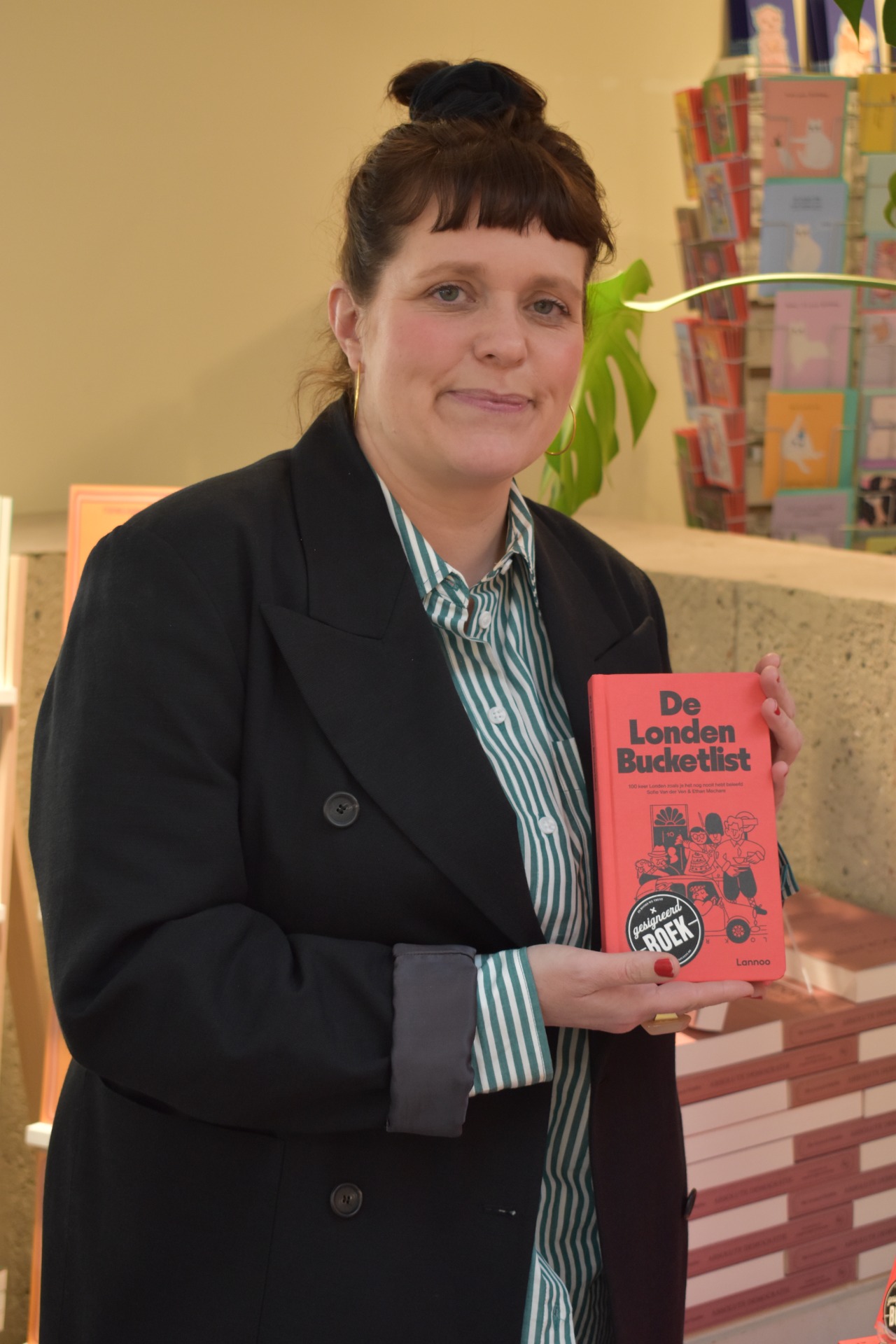 Vrouw in blazer houdt rood boek “De Londen Bucketlist” vast, met boekenplanken en plant op achtergrond.