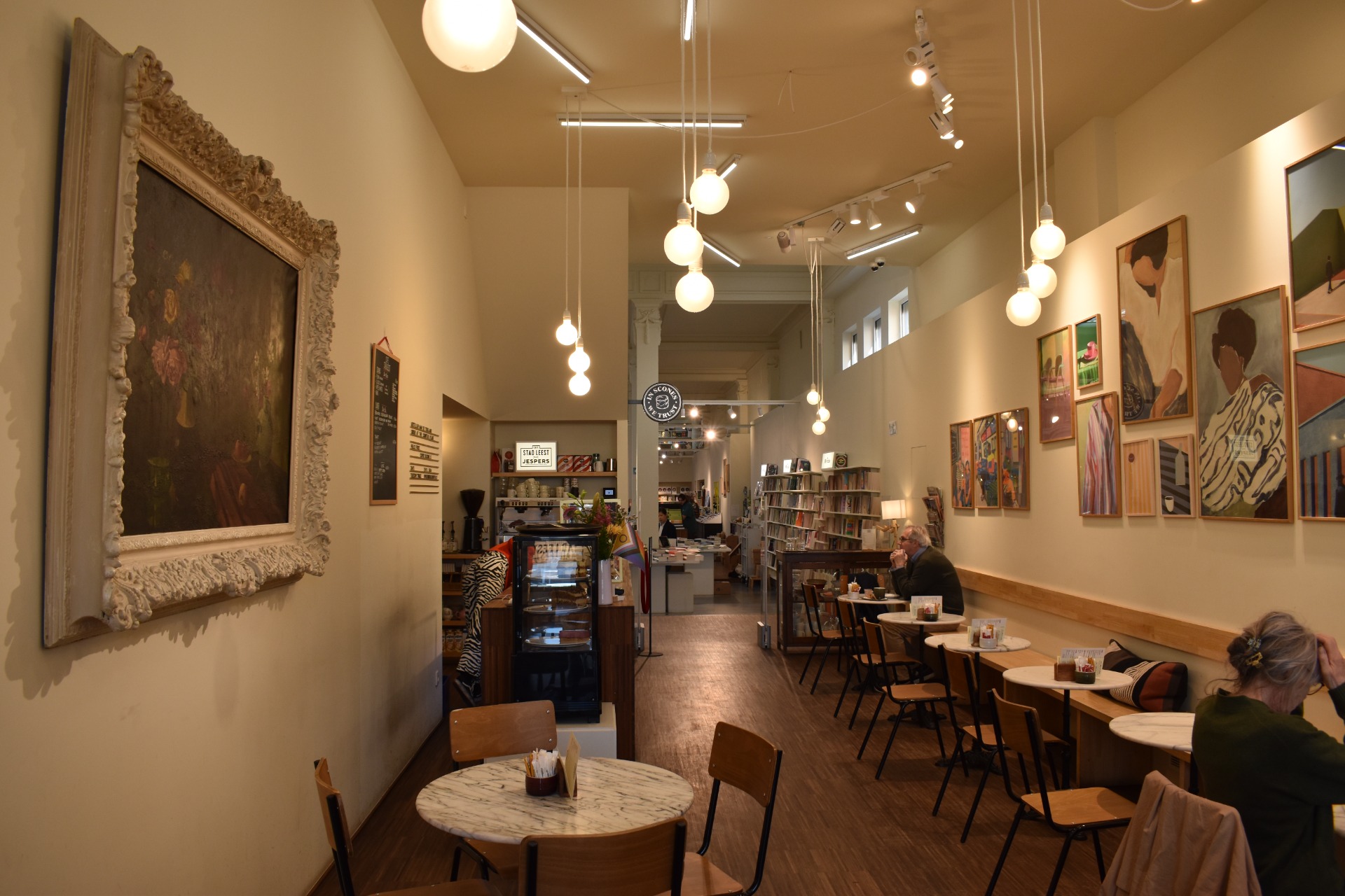 Gezellige galerie-koffiezaak met kunst aan muren, tafels, glazen vitrine en hangende lampen in lange gang.