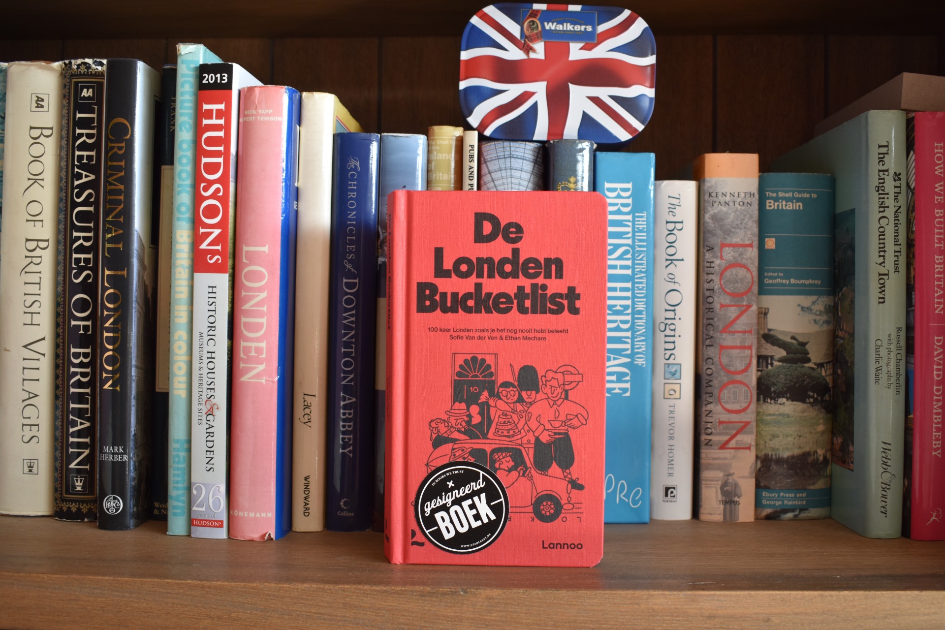 Boekenkast met diverse boeken; vooraan staat rood boek “De London Bucketlist” met illustraties.