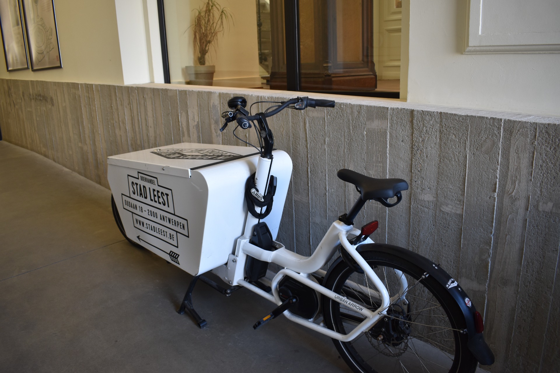 Witte deelfiets met zwarte stuur en zadel, geparkeerd tegen stenen muur in gang met ramen.