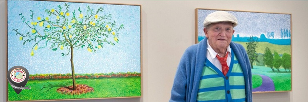 David Hockney staat voor twee ingelijste schilderijen met een boom en een landschap aan de muur.