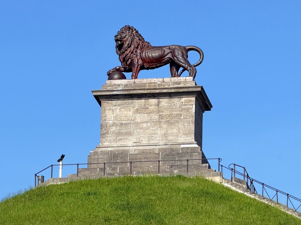 Bronzen leeuw standbeeld op stenen sokkel met inscriptie, op groene heuvel tegen blauwe lucht.