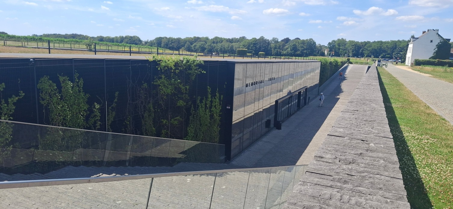 Langgerekte betonnen muur met opschrift, pad met bezoekers en groene velden onder blauwe lucht.