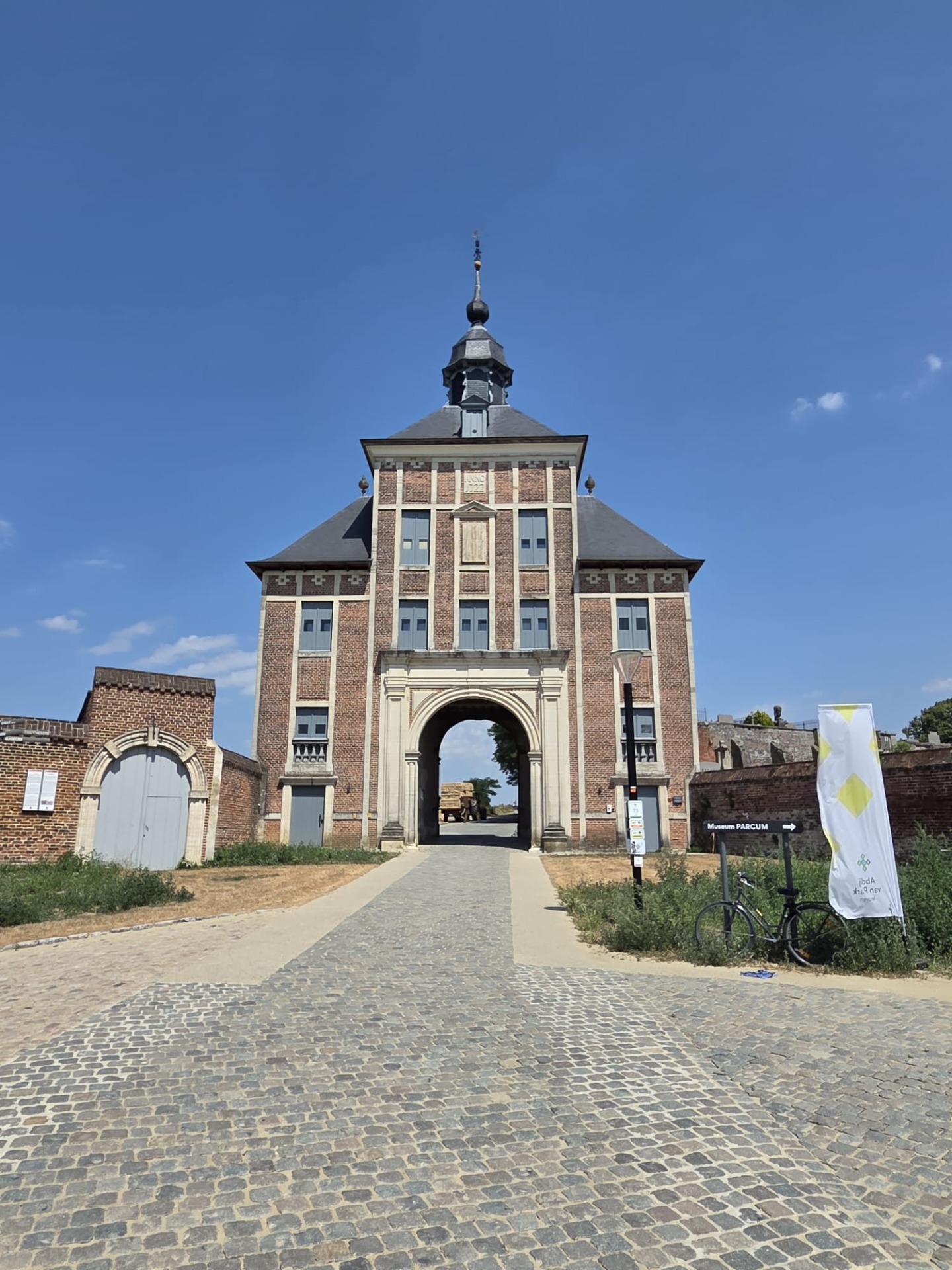 Bakstenen poortgebouw met boogdoorgang, klokkentoren en vlaggen, gezien vanaf een geplaveide oprijlaan onder blauwe lucht.