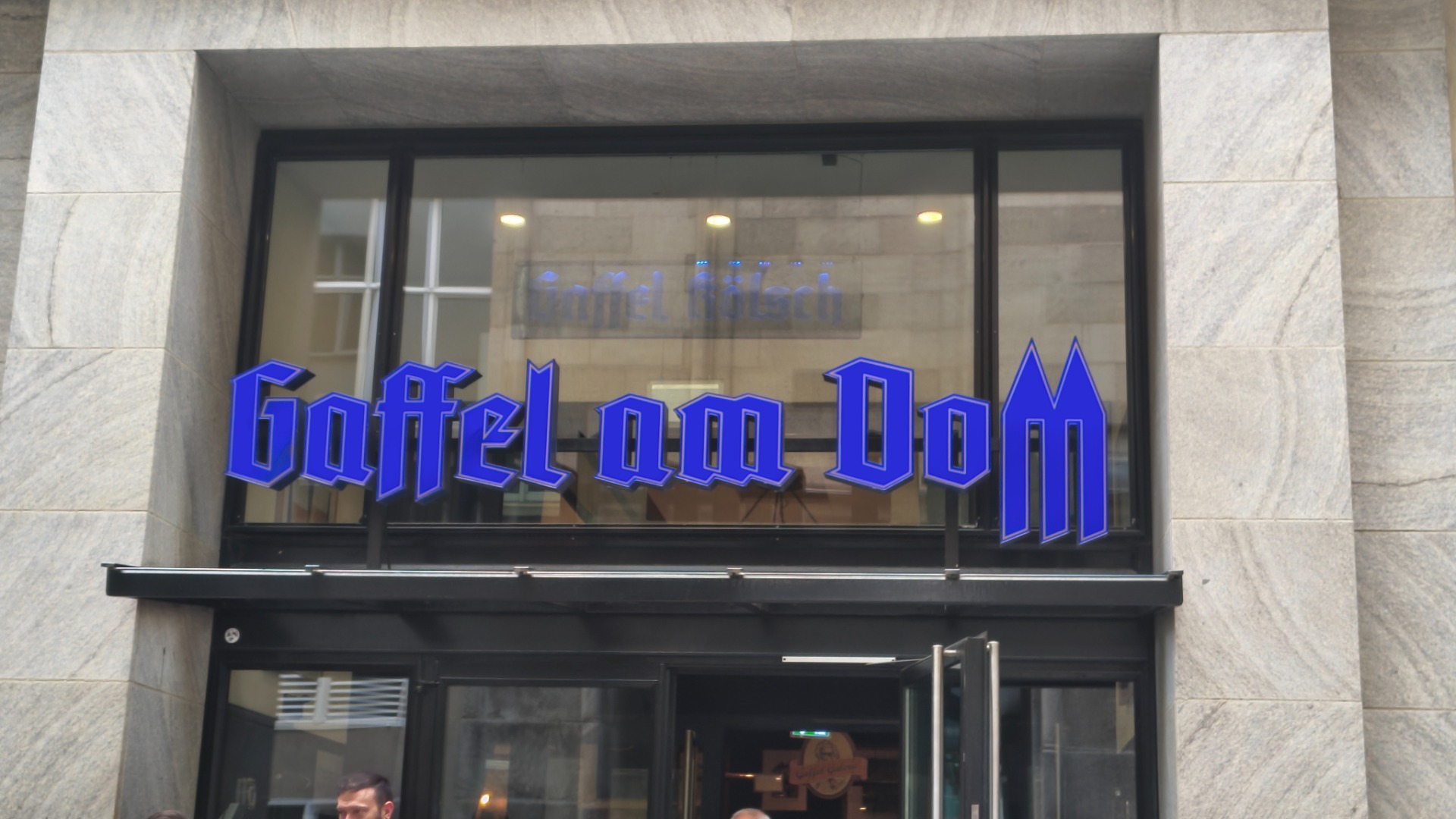 Blauwe neonletters “Gaffel Am Dom” op glazen gevel, binnenverlichting en stenen muur rondom.