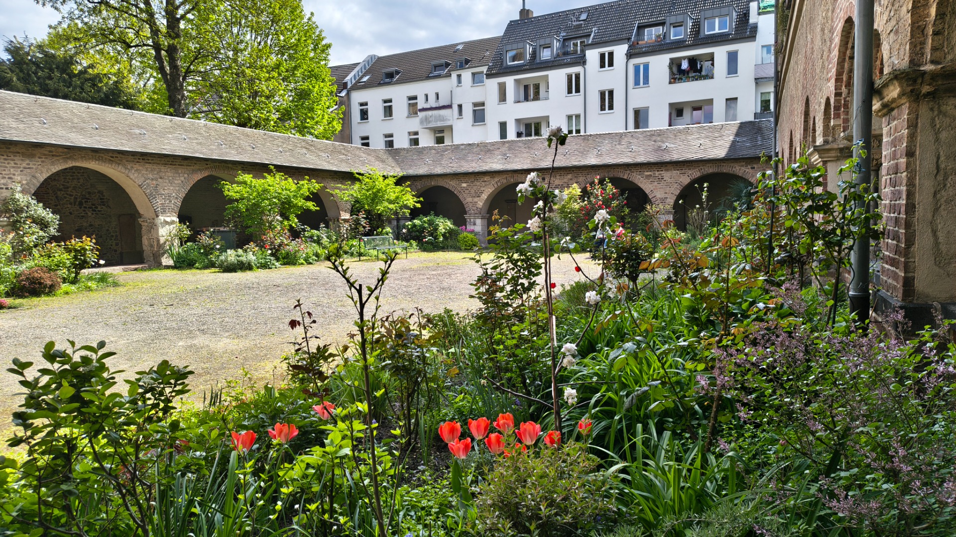 Bloementuin met rode tulpen en groene planten, binnenplaats met stenen bogen en witte appartementen op achtergrond.