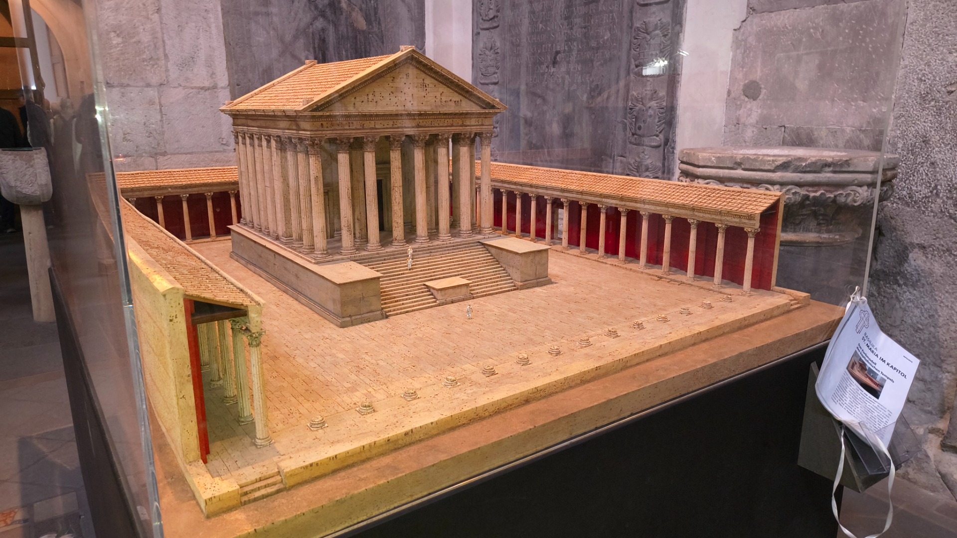 Miniatuur van een Romeins tempelcomplex met zuilen, rood interieur en trap, tentoongesteld in museumruimte.