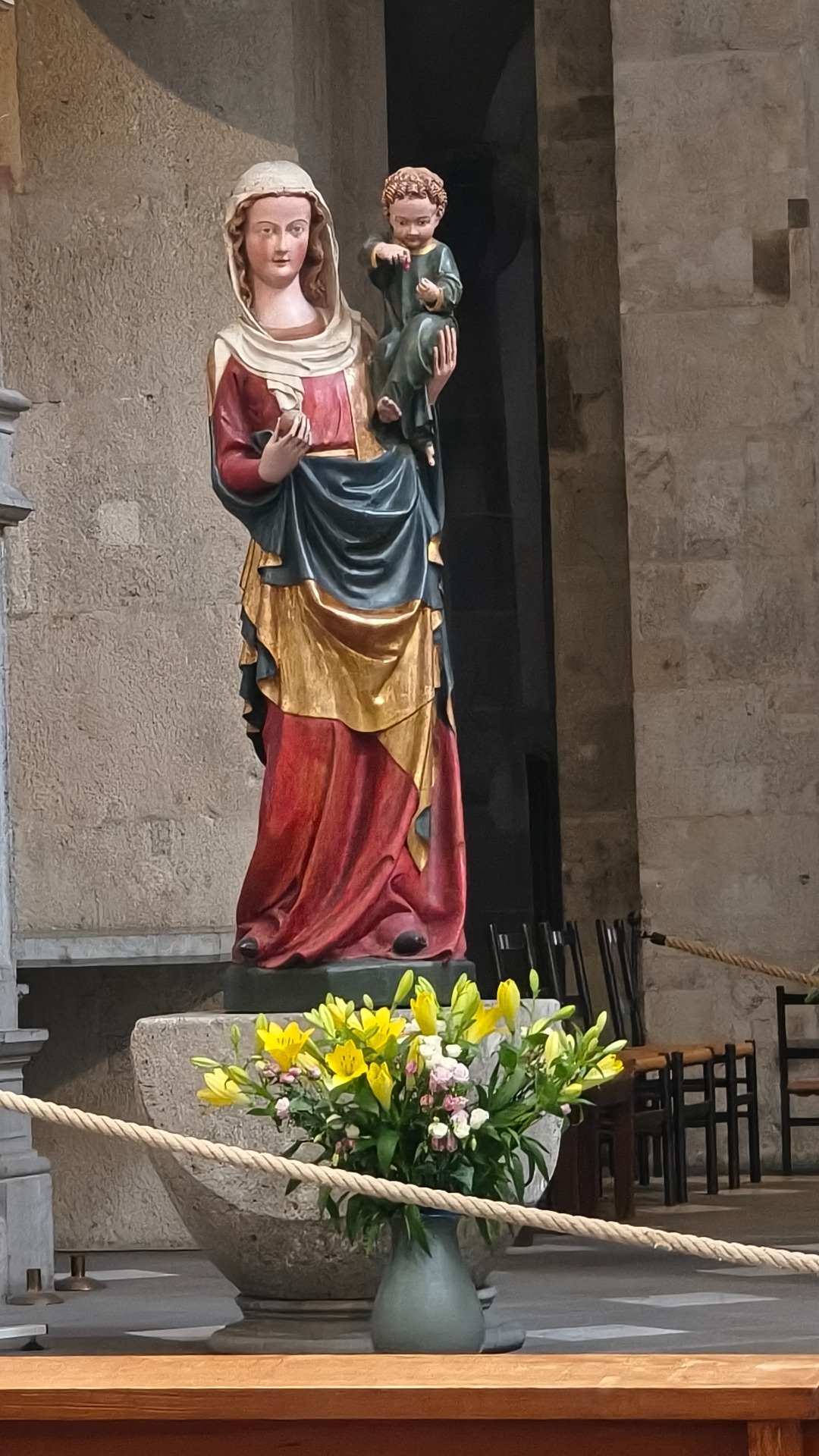 Gekleurde heiligenbeelden van Maria en kind Jezus, met bloemenboeket op altaar in stenen kerk.