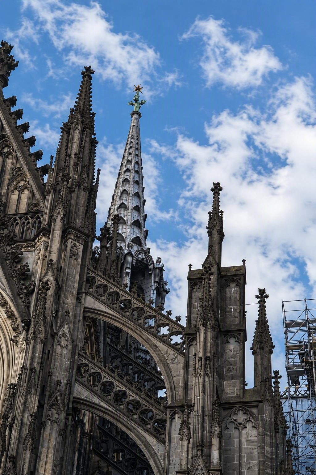 Gotische kathedraal met hoge torens, bogen en steigers, tegen een blauwe lucht met wolken.