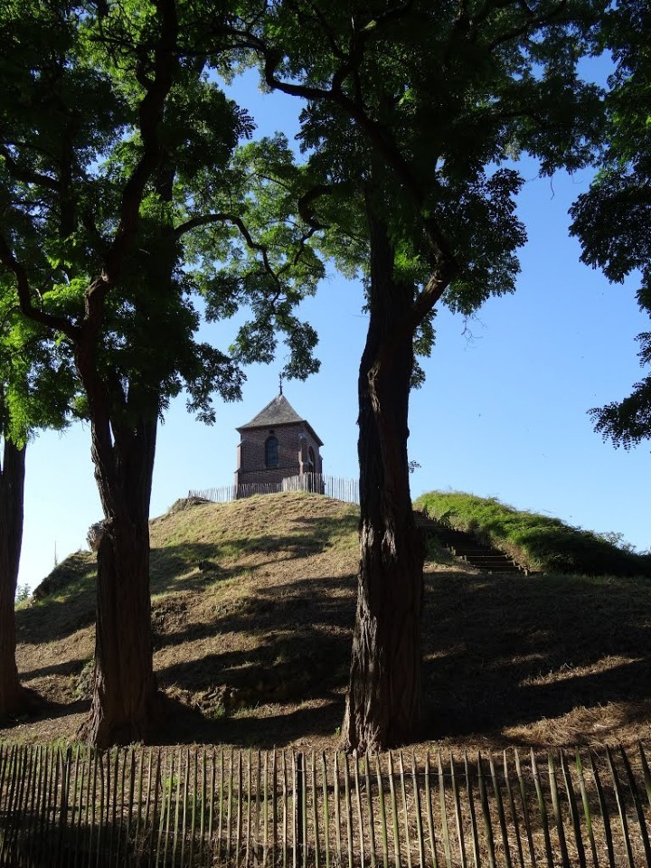 Kapelletje op heuvel met trap, omringd door hoge bomen en een houten hek, blauwe lucht.