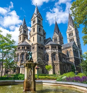 Grote gotische kathedraal met torenspitsen, fontein en vijver, omringd door bomen en blauwe lucht met wolken.