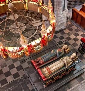 Kerkinterieur met ronde kroonluchter, stenen zuilen en opgevouwen tapijten op een zwarte en witte tegelvloer.