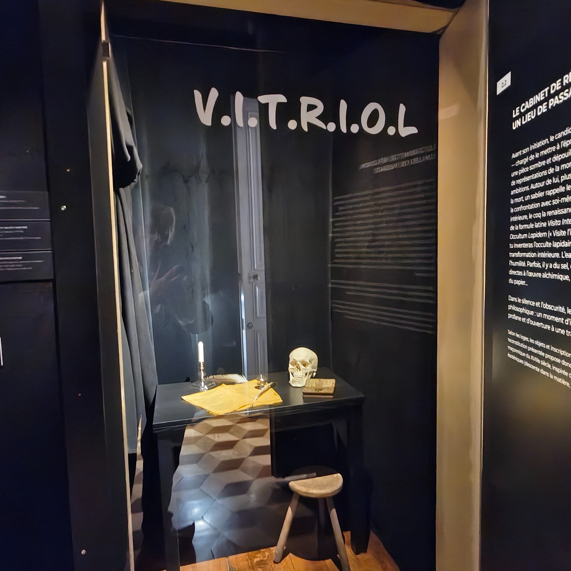 Tentoonstellingsvitrine met schedel, kaars en papieren op tafel, met tekst “V.IT.R.I.O.L” op zwarte wand.
