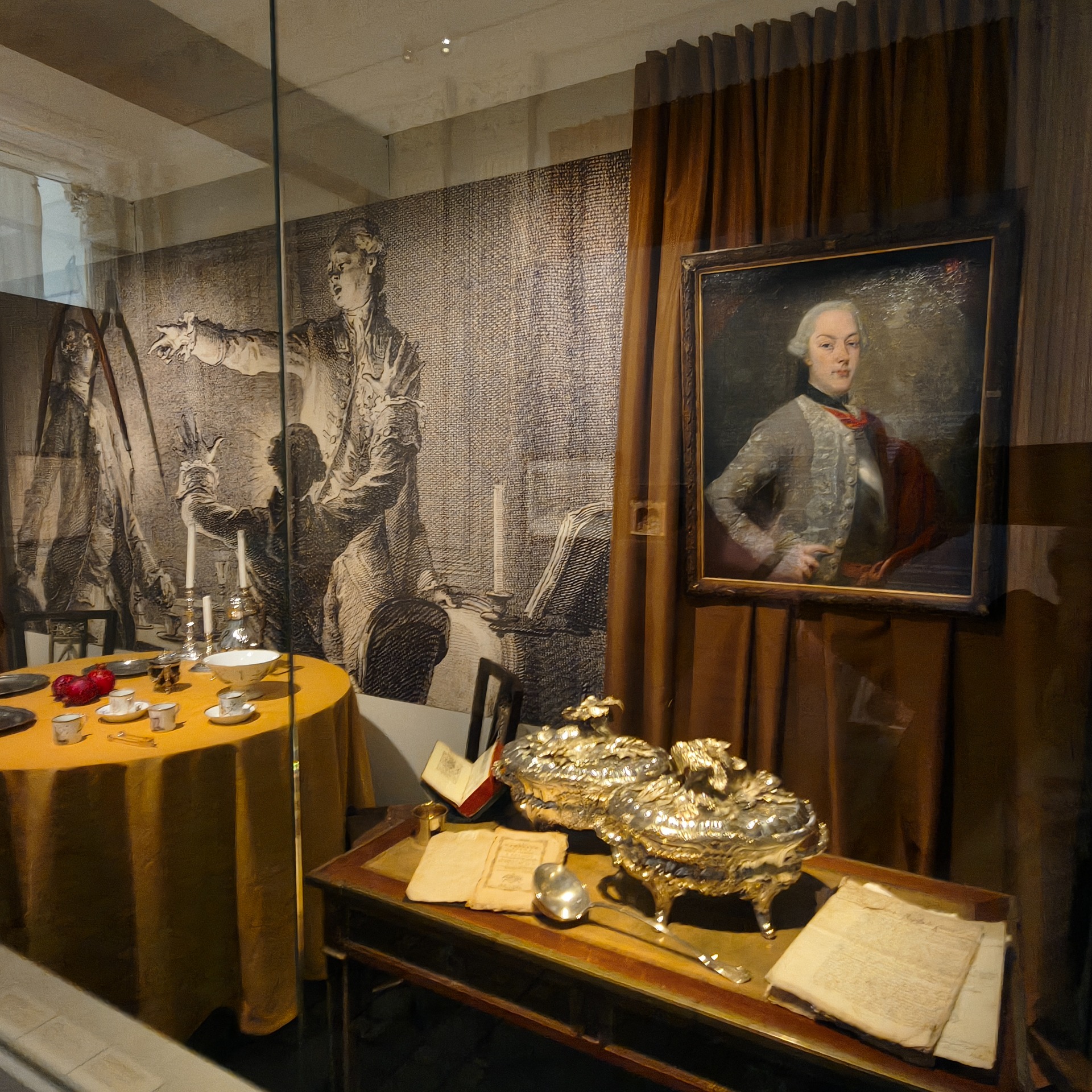 Tentoonstellingsruimte met schilderijen, een gedekte tafel en kandelaars, met een man in portret op doek.