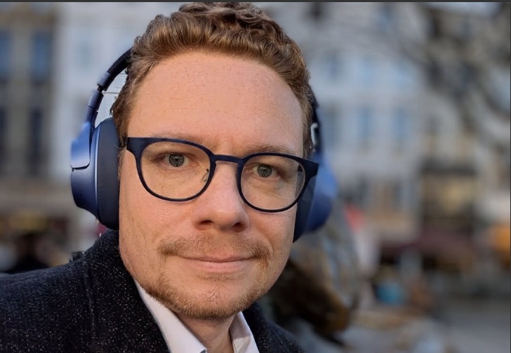 Man met grote bril en blauwe koptelefoon, close-up voor onscherpe stadsachtergrond.