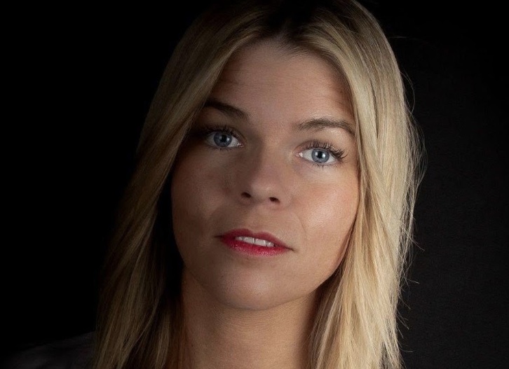 Portret van een blonde vrouw met blauwe ogen, neutrale blik en zachte belichting tegen donkere achtergrond.