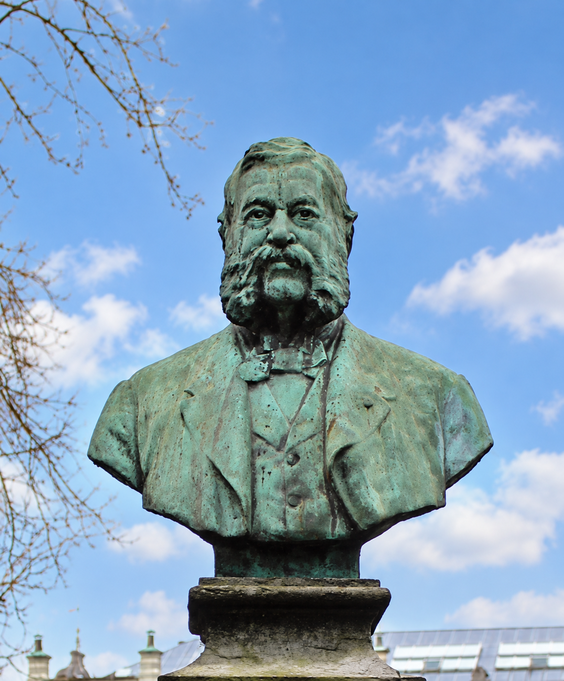 Groene bronzen borstbeeld van een man met snor en stropdas tegen blauwe lucht met wolken.