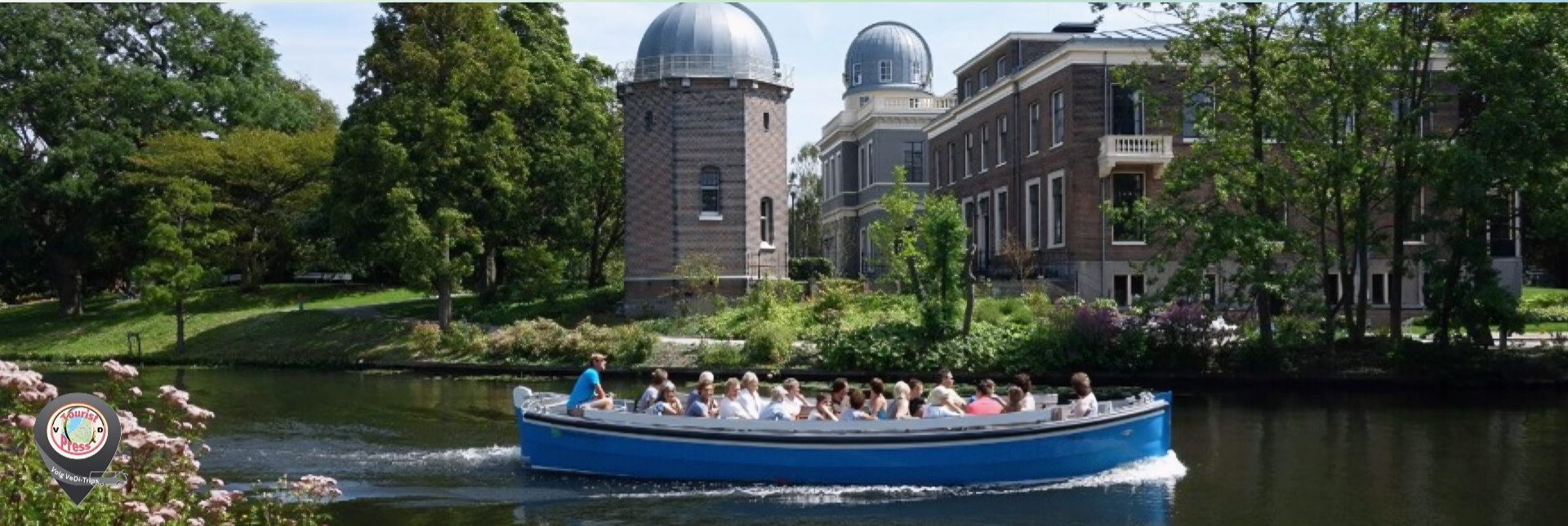 Blauwe rondvaartboot met passagiers vaart op vijver voor historische gebouwen met koepels en bomen.