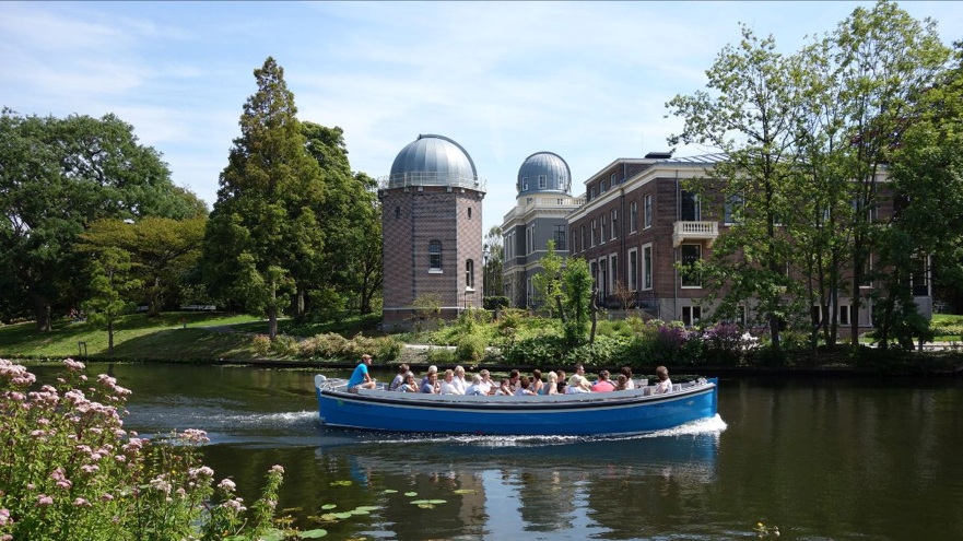 Blauwe rondvaartboot met passagiers op een vijver, met bakstenen toren en koepelgebouwen op de achtergrond.