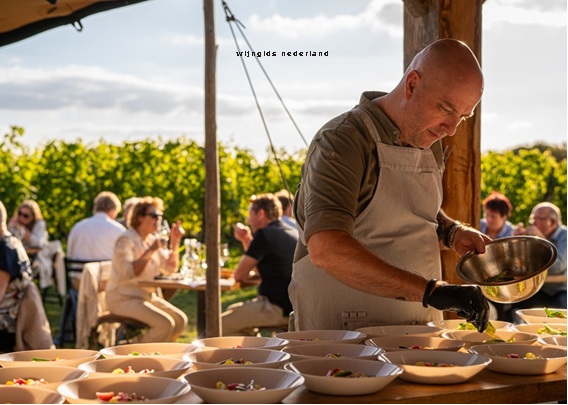 Kok schept eten in kommen tijdens buiten catering, terwijl gasten op terras in de achtergrond kijken.