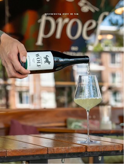 Hand schenkt witte wijn uit fles in glas op houten bar, achtergrond met “proef” en terrasdecor.