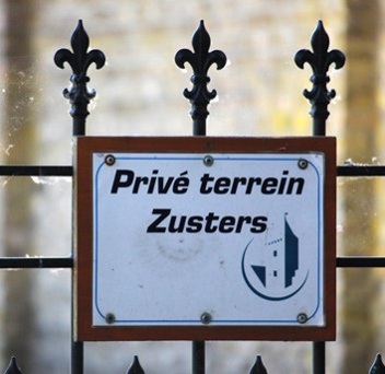 Bord met tekst “Privé terrein Zusters” tussen smeedijzeren hekwerk met fleur-de-lis ornamenten.